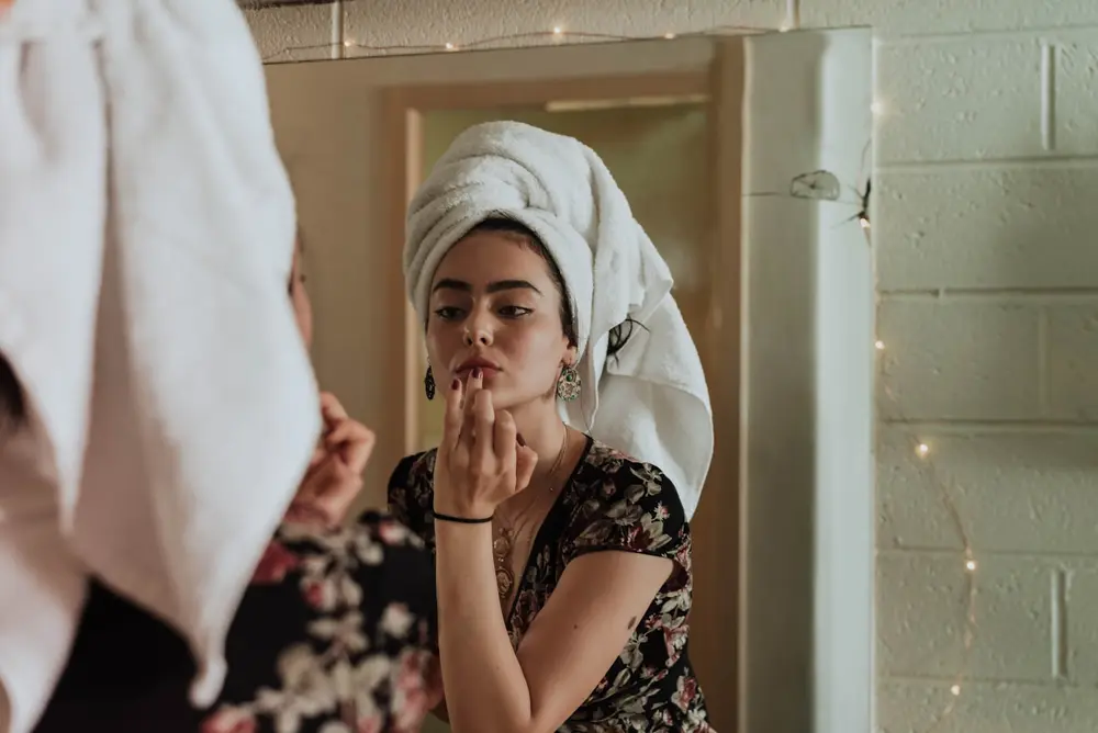 Woman with a towel wrapped in her hair going through her morning skincare routine at the mirror